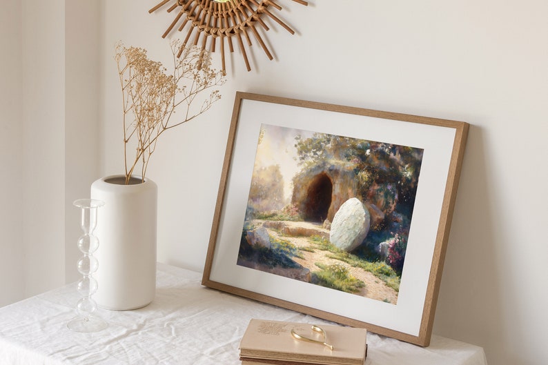 May include: A framed painting depicting a stone tomb with a large, round stone rolled away from the entrance. The tomb is surrounded by lush greenery and flowers, suggesting a peaceful and hopeful scene.