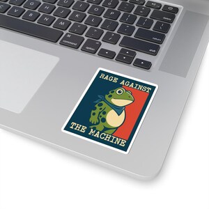 Op de afbeelding: Een sticker met een cartoonkikker met groene stippen, een blauwe bandana en de tekst "RAGE AGAINST THE MACHINE". De kikker staat tegen een rode en blauwe achtergrond, met witte randen.