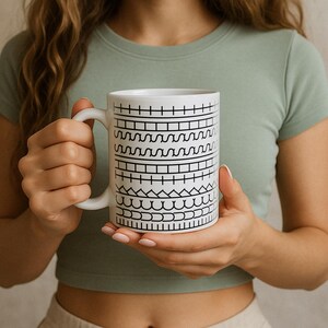 May include: White ceramic mug with a black geometric pattern. The mug features a series of horizontal bands with different black line designs. The mug has a white handle and is held by a person wearing a green shirt.