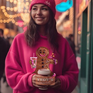 May include: A bright pink hooded sweatshirt featuring a graphic of a gingerbread person holding a cup with a straw. The gingerbread person has a smiling face, three dark buttons, and pink icing accents. The sweatshirt is paired with a matching pink beanie.
