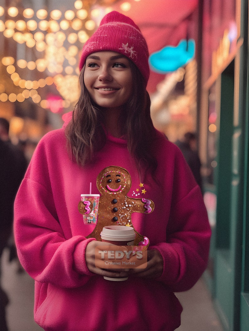 May include: A bright pink hooded sweatshirt featuring a graphic of a gingerbread person holding a cup with a straw. The gingerbread person has a smiling face, three dark buttons, and pink icing accents. The sweatshirt is paired with a matching pink beanie.