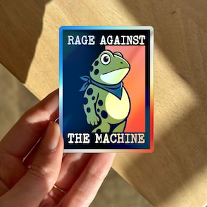 Op de afbeelding: Holografische sticker met een cartoon kikker met een blauwe bandana. De tekst op de sticker luidt "RAGE AGAINST THE MACHINE". De kikker is groen met zwarte stippen, tegen een gradi&euml;nt achtergrond.