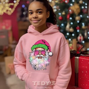 Op de afbeelding: Een lichtroze hoodie met een afbeelding van de kerstman met een zonnebril en een kleurrijke baard. De afbeelding van de kerstman staat op de voorkant. De hoodie heeft een voorzak en de tekst "TEDY'S Creative Market" is eronder gedrukt.