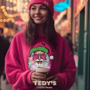 Op de afbeelding: Een vrouw draagt een roze beanie en een roze hoodie met een kerstman-afbeelding. De kerstman heeft een zonnebril en kleurrijke lampjes. De tekst "TEDY'S Creative Market" staat op de hoodie.