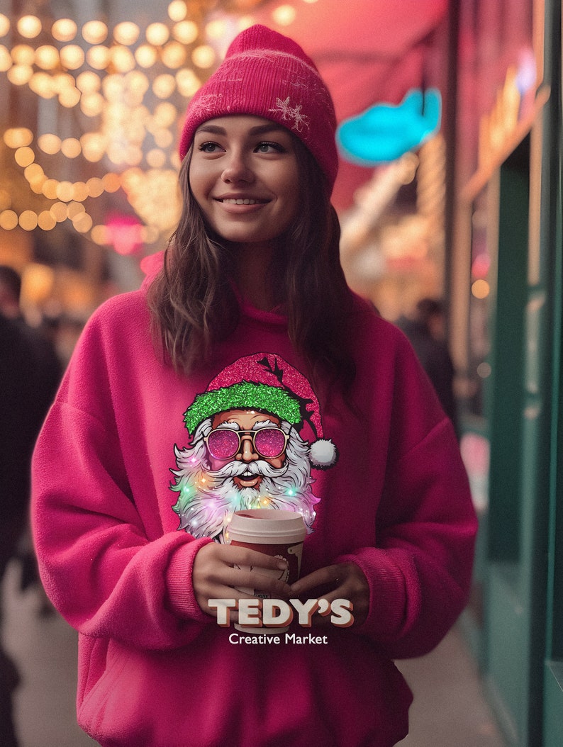 Op de afbeelding: Een vrouw draagt een roze beanie en een roze hoodie met een kerstman-afbeelding. De kerstman heeft een zonnebril en kleurrijke lampjes. De tekst "TEDY'S Creative Market" staat op de hoodie.