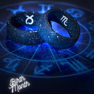 May include: Two dark blue rings with white speckles and zodiac symbols. One ring displays the Taurus symbol, the other the Scorpio symbol. The rings are set against a blue background with a zodiac chart and the words "Birth Month".