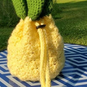May include: A handmade crochet pineapple-shaped bag. The bag is yellow with a drawstring closure and green leaves at the top. The bag is sitting on a blue and white geometric patterned surface.