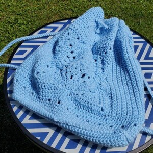 May include: A light blue crochet drawstring bag with a textured floral design on the front. The bag has a ribbed knit pattern on the sides and a matching drawstring closure. The bag is sitting on a blue and white patterned table.