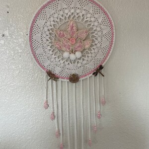 May include: White crochet dreamcatcher with a pink flower design and pink beads hanging from the bottom.