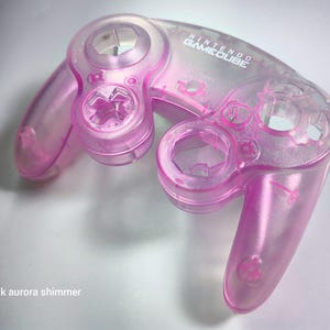 May include: A translucent pink Nintendo GameCube controller with a shimmery finish. The controller has a clear plastic design and the text "NINTENDO GAMECUBE" is printed on the top.