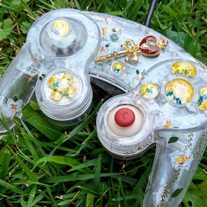 May include: A clear, translucent GameCube controller with a red button and gold accents. The controller is decorated with white flowers and gold glitter.