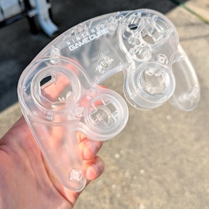 May include: A clear, transparent Nintendo GameCube controller. The controller has a classic design with buttons, a directional pad, and analog sticks. The text "NINTENDO GAMECUBE" is visible on the top. The controller is held in a hand.