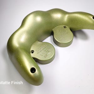 May include: A metallic olive green controller with a matte finish. The controller has a curved, ergonomic design with two circular buttons and two holes. The text "Matte Finish" is visible at the bottom.