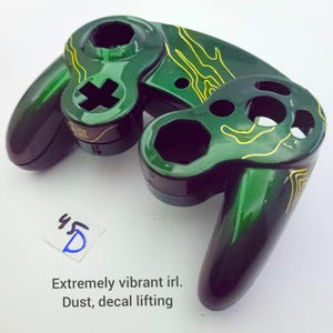 Puede incluir: Controlador de GameCube verde y dorado con una pintura personalizada y una calcomanía. La calcomanía tiene un patrón de ondas y se está despegando en algunas áreas. El controlador está polvoriento.