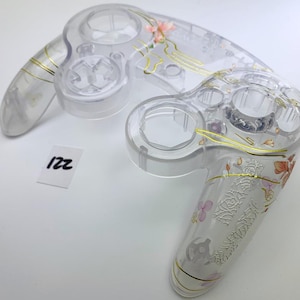 May include: A clear plastic GameCube controller shell with a floral design and gold accents. The shell is translucent and shows the internal components of the controller.