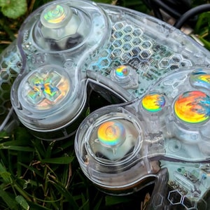 May include: A clear plastic GameCube controller with iridescent buttons. The controller has a honeycomb pattern on the back and is lying on green grass.