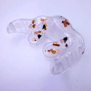May include: A clear plastic mold shaped like a hand with a textured surface. The mold is filled with small, colorful confetti.