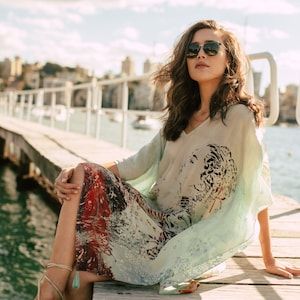 May include: A woman wearing sunglasses sits on a wooden jetty wearing a light blue and white patterned kimono with red and black accents. The kimono has a tiger print design.