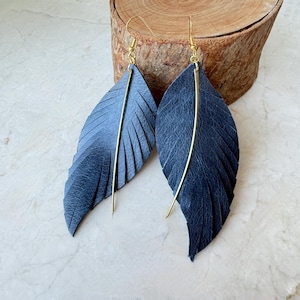 Puede incluir: Un par de pendientes de pluma de cuero azul marino con alambres para las orejas dorados y un acento vertical dorado. Los pendientes se exhiben sobre una superficie clara con un fondo de madera.
