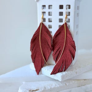 May include: A pair of burgundy feather earrings with gold-colored accents. The earrings are made of a soft, textured material and feature a long, slender gold bar down the center. The earrings are displayed on a white surface.