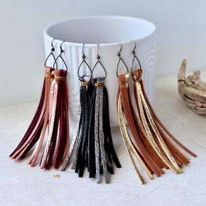 May include: Five pairs of tassel earrings in a variety of colours. The earrings have long, thin leather tassels in burgundy, black, silver, brown, and gold. Each pair has a unique design, with metal accents and ear wires.