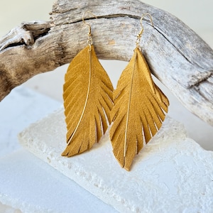 Puede incluir: Un par de pendientes de plumas de color amarillo mostaza con ganchos dorados y una fina línea dorada. Los pendientes se exhiben sobre un fondo neutro con un trozo de madera flotante.