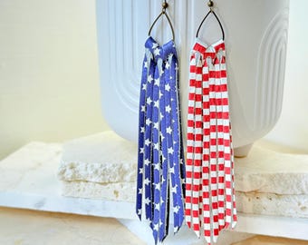 May include: A pair of patriotic earrings. One earring features a blue and white star pattern, while the other has a red and white checkered design. Both earrings have bronze-colored ear wires and a fringe-like design.