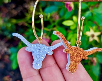 May include: A pair of gold hoop earrings with a glitter-covered Texas Longhorn head charm on each earring. One charm is white and the other is orange.