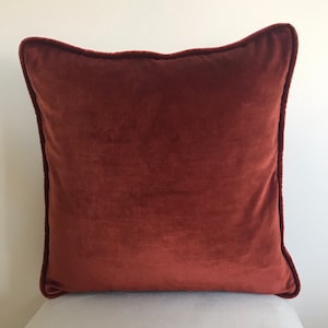 May include: A rust-colored velvet pillow with a simple, square shape and a subtle sheen. The pillow is sitting on a white surface.