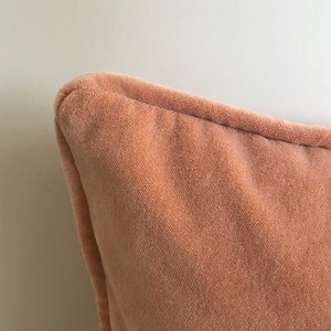 May include: Close-up of a peach-colored velvet pillow with a subtle texture. The pillow is soft and plush, with a smooth, even surface.
