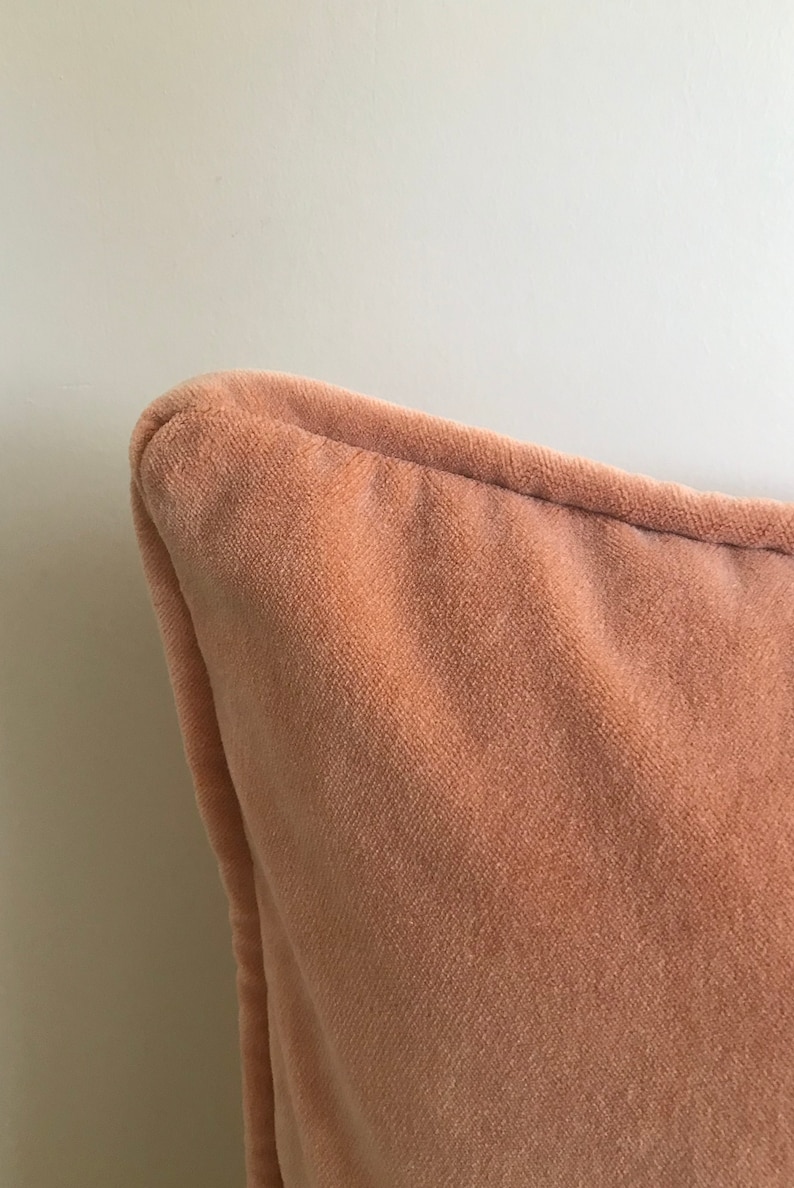 May include: Close-up of a peach-colored velvet pillow with a subtle texture. The pillow is soft and plush, with a smooth, even surface.