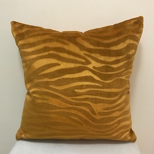 May include: Mustard-colored decorative pillow with a zebra stripe pattern. The pillow has a soft, velvety texture and a square shape. The zebra stripes are a lighter shade of mustard, creating a contrasting design.