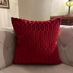 May include: A red velvet throw pillow with a geometric pattern. The pillow is on a white couch.