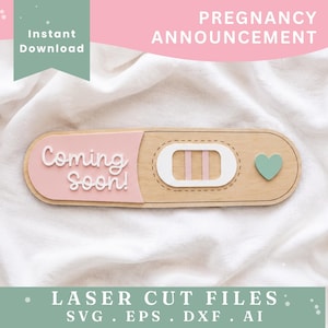 May include: A wooden pregnancy test prop featuring a light pink panel with the white text Coming Soon!, a white rectangular window with two vertical pink lines, and a small light green heart accent on a light wood base with rounded edges.