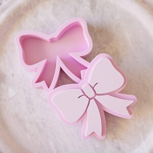 May include: Two light pink bow-shaped cookie cutters. One cutter is an open frame outline, while the other is a solid-faced stamp with embossed details showing the center knot and ribbon tails of a bow. Both items are made of smooth plastic.