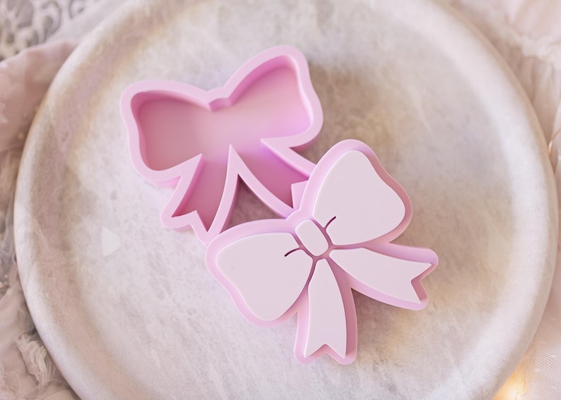 May include: Two light pink bow-shaped cookie cutters. One cutter is an open frame outline, while the other is a solid-faced stamp with embossed details showing the center knot and ribbon tails of a bow. Both items are made of smooth plastic.