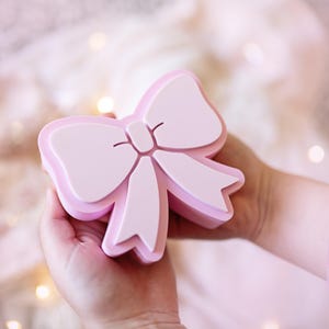 May include: A light pink, three-dimensional bow-shaped object with a smooth, matte finish. The design features a central knot with two loops and two downward-hanging ribbon tails, all rendered in a solid, uniform pastel pink color.