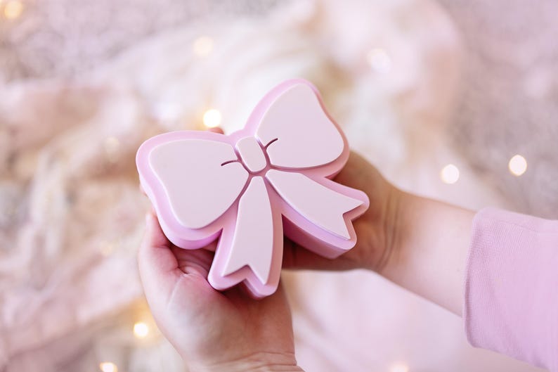 May include: A light pink, three-dimensional bow-shaped object with a smooth, matte finish. The design features a central knot with two loops and two downward-hanging ribbon tails, all rendered in a solid, uniform pastel pink color.