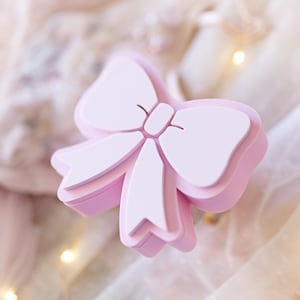 May include: A light pink, three-dimensional bow-shaped object with a smooth, matte finish. The design features a central knot with two loops and two downward-pointing ribbon tails, creating a layered, structured appearance with clean edges.