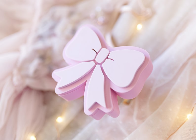 May include: A light pink, three-dimensional bow-shaped object with a smooth, matte finish. The design features a central knot with two loops and two downward-pointing ribbon tails, creating a layered, structured appearance with clean edges.