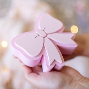 May include: A light pink, bow-shaped box with a smooth, matte finish. The lid features a raised, stylized bow design that matches the base. The container is solid in color and has a clean, simple structure with rounded edges and a distinct two-part design.