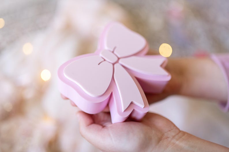 May include: A light pink, bow-shaped box with a smooth, matte finish. The lid features a raised, stylized bow design that matches the base. The container is solid in color and has a clean, simple structure with rounded edges and a distinct two-part design.