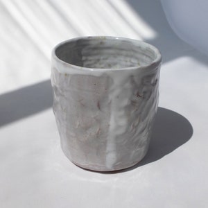 May include: A handmade, off-white ceramic cup with a textured, uneven surface. The cup has a cylindrical shape with a slightly flared rim. The exterior features a rough, organic texture, and the interior is smooth. The cup is suitable for beverages.