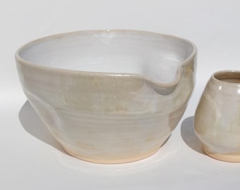 May include: A ceramic mixing bowl with a wide, rounded shape and a small pouring spout, featuring a glossy, off-white and beige glaze with a smooth finish. Beside it sits a smaller, matching ceramic vessel with a tapered, rounded body.