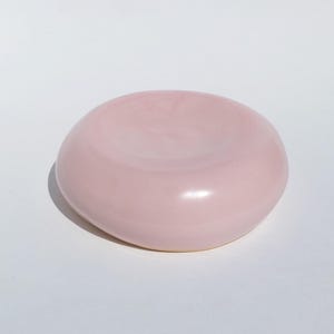 May include: A pale pink ceramic bowl with a smooth, glossy surface. The bowl has a shallow, curved interior and a slightly raised edge. The design is organic and contemporary, suitable for serving or decorative purposes.