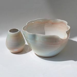 May include: A ceramic set consisting of a small, pear-shaped vase and a larger, wide-mouthed bowl with a scalloped rim and a pouring spout. Both pieces feature a smooth, glossy finish with a soft, iridescent blend of pastel pink, blue, and white.