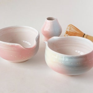 May include: A collection of pastel-colored ceramic tea ceremony items. The set includes two bowls with spouts, a small vase, and a bamboo whisk. The bowls and vase feature a gradient of pink, blue, and white. The whisk is made of thin bamboo.