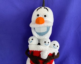 May include: A white crocheted snowman figure with a carrot-shaped nose, black twig-like hair, and dark brown arms. The snowman holds a red rectangular pouch containing three small white snow-creature figures with blue smiles and black eyes.