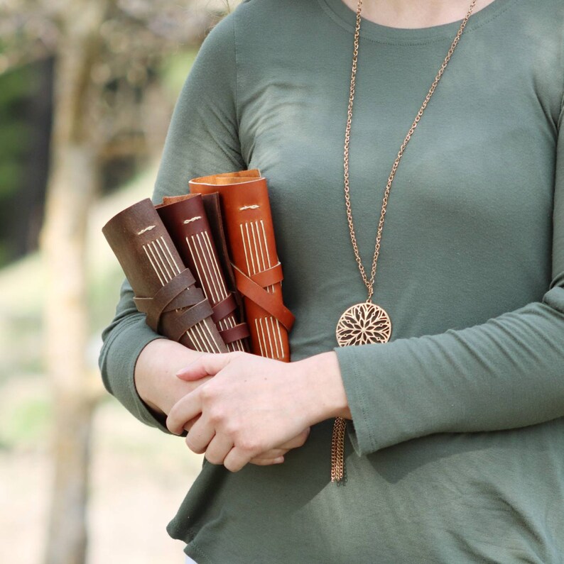 May include: Three leather-bound journals with decorative stitching and a brown, tan, and orange colour scheme. The journals are held in the arms of a person wearing a green shirt and a gold necklace with a circular pendant.