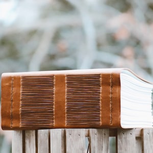 May include: A brown leather journal with a stitched spine and a series of horizontal lines on the cover. The pages are thick and white. The journal is resting on a wooden surface. The notebook is likely for writing or sketching.